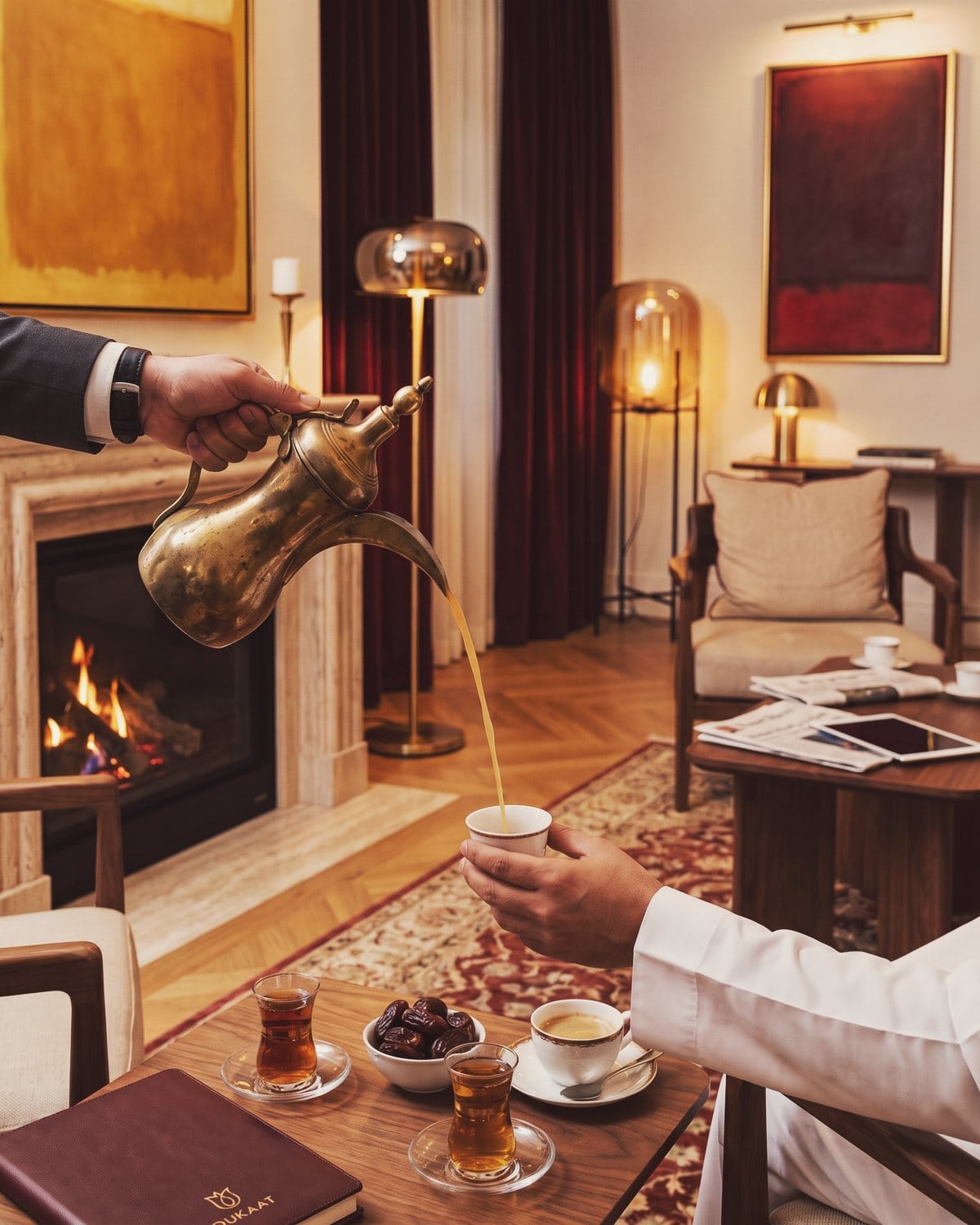 Arabic coffee is poured from a brass dallah beside tea glasses and dates by the fireplace.